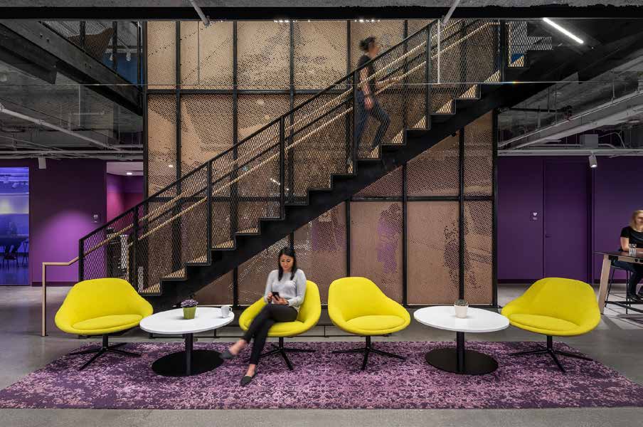An open-plan office where a black steel stair climbs a wire-mesh screen wall, with yellow lounge chairs and round white tables gathered on a patterned rug below.