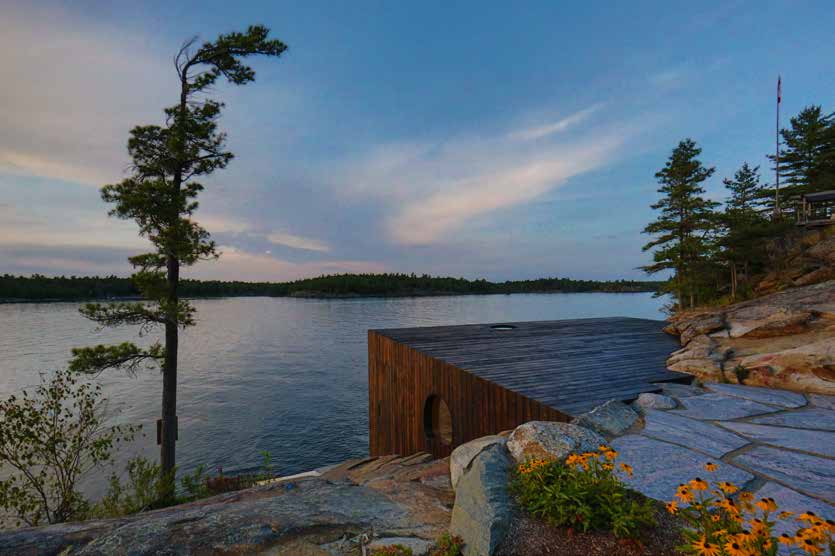 The charred cedar volume set low on a pink granite shore at dusk, its oval window facing a calm bay, with a lone windswept pine and black-eyed Susans in the foreground.