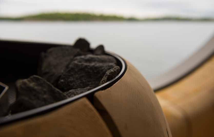 A close view of a black sauna stove filled with dark rocks, set into the pale curved cedar floor, with the lake and a wooded shoreline soft behind it.