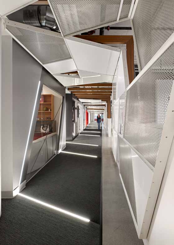 A corridor of angled white panels and glazed partitions narrowing toward a distant figure, lit by a continuous recessed strip.
